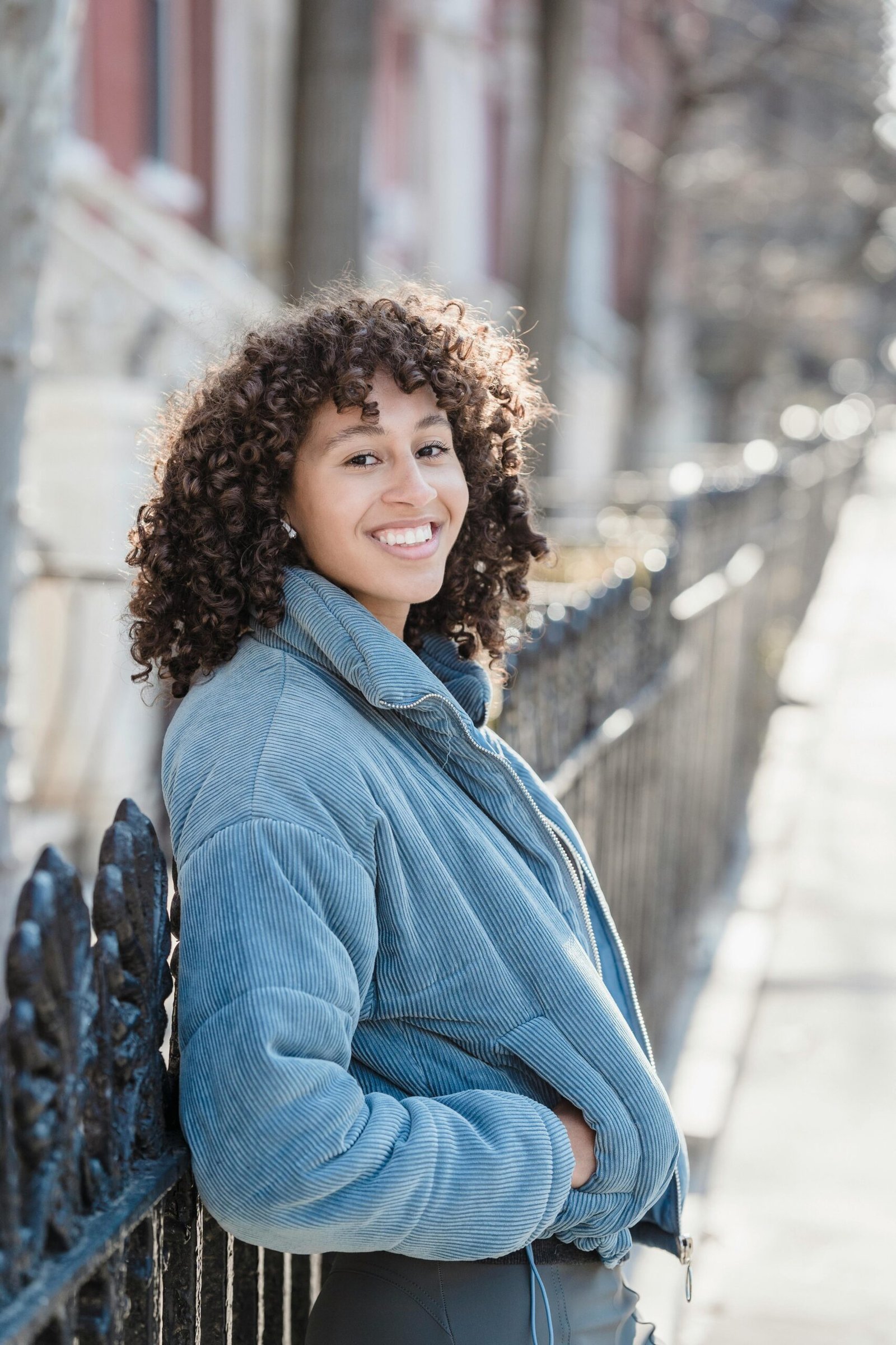 Smiling woman with curly hair leaning against a fence outdoors, enjoying a sunny day.